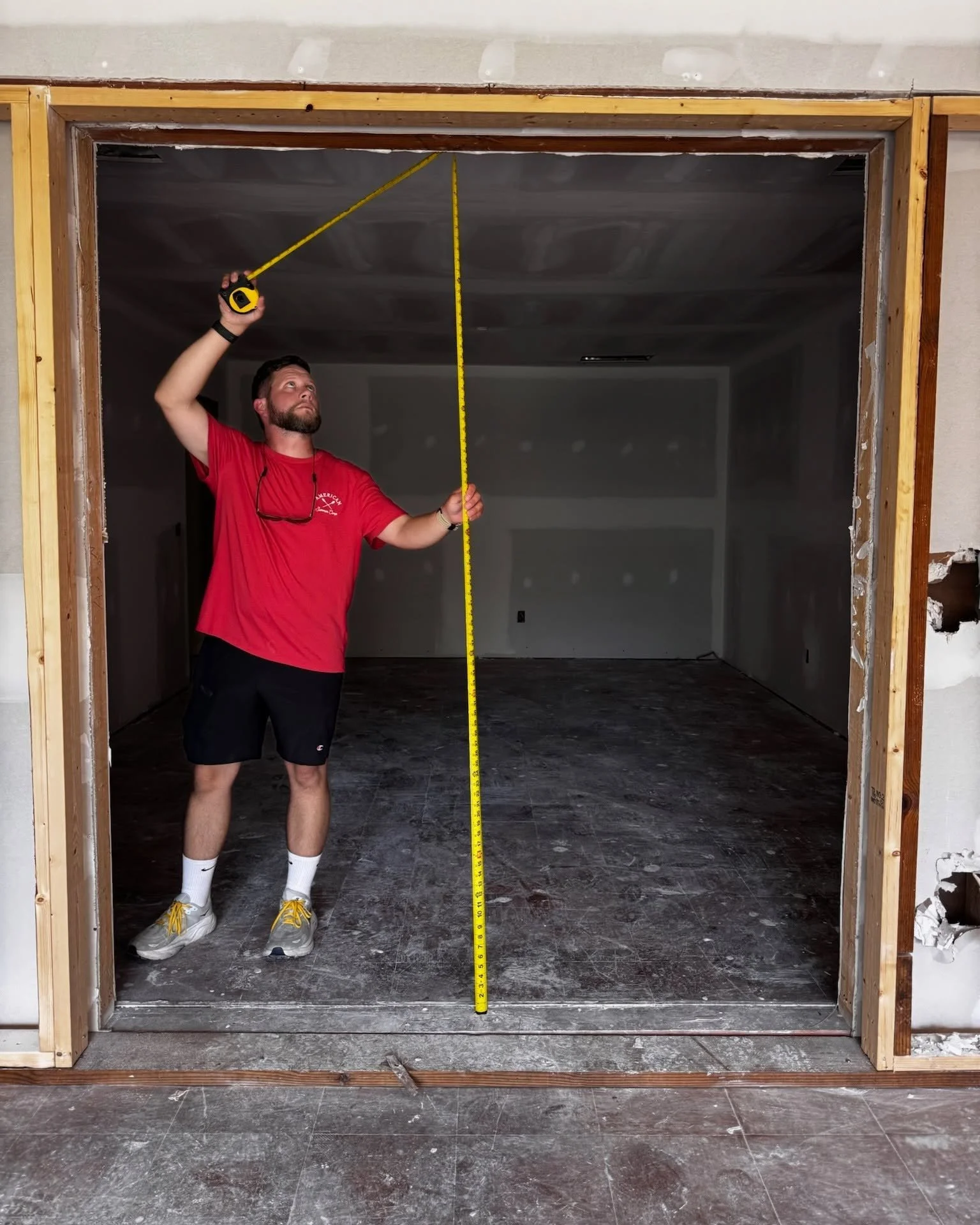 A man in a red t-shirt and black shorts is measuring the height of a doorframe with a tape measure, standing inside an unfinished room with exposed wall studs and drywall interrupts on the side.