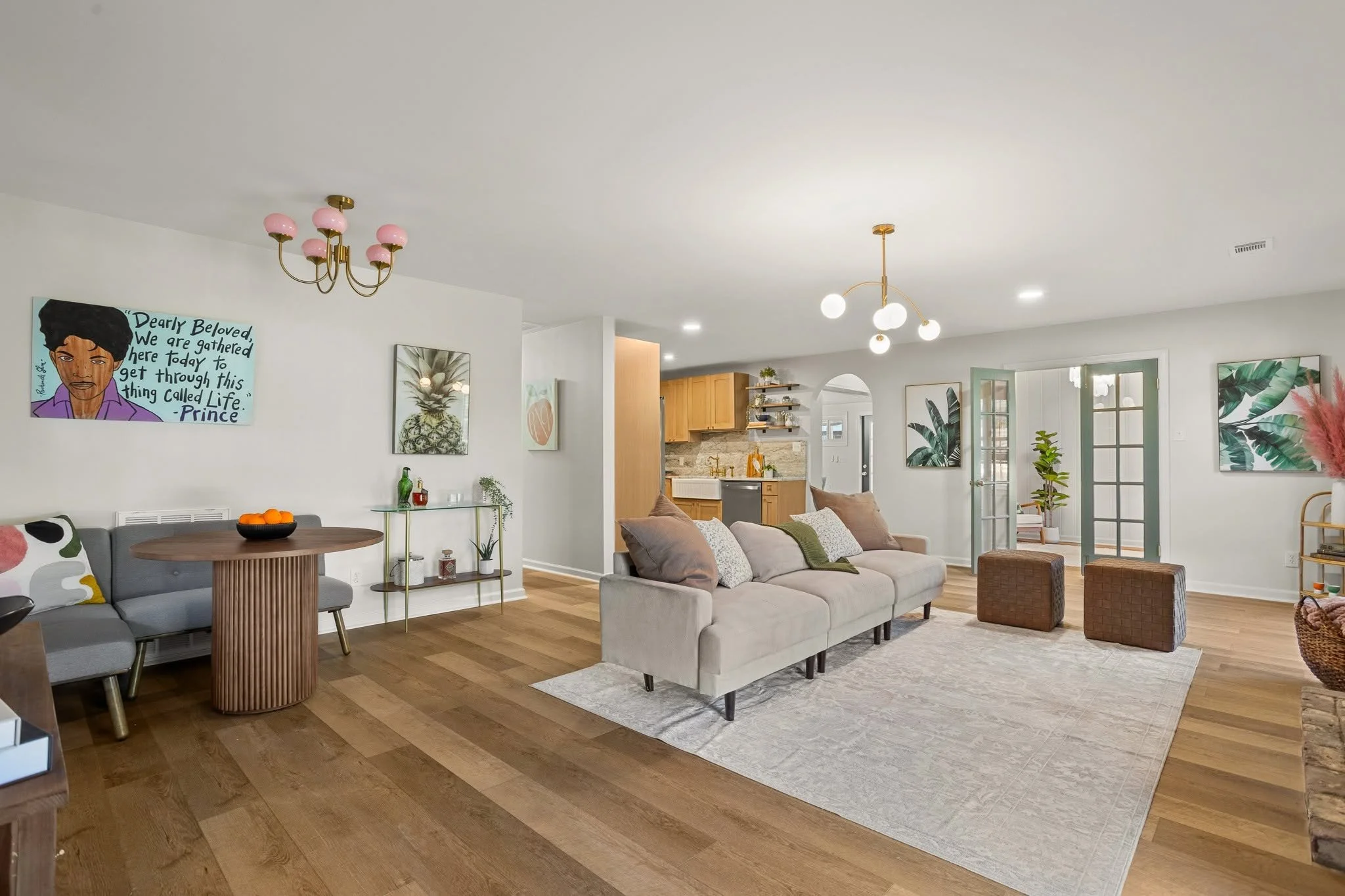 Open-concept living room with a light-colored sofa, patterned cushions, and a white area rug. Decor includes framed artwork featuring a pineapple, tropical leaves, and a face with a quote. There is a modern dining table with chairs, surrounded by wal