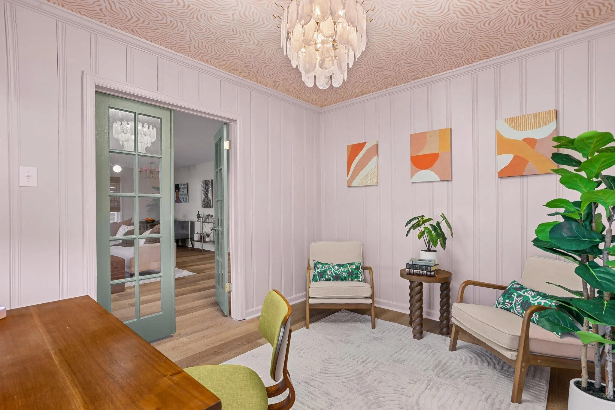 A cozy living room with beige walls and wooden floors. There are two chairs with patterned cushions and a small round side table with books. A large potted plant sits in the corner. Abstract artwork decorates the wall. A door with glass panes leads t