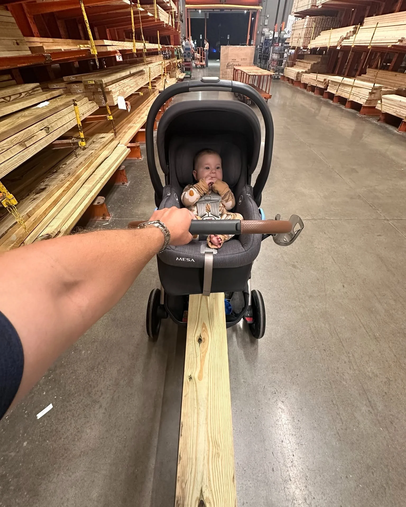 A baby sitting in a stroller in a hardware store aisle with wood planks on the shelves. An adult's hand is holding the stroller's handle, and there is a wooden plank on the floor aligned with the stroller.