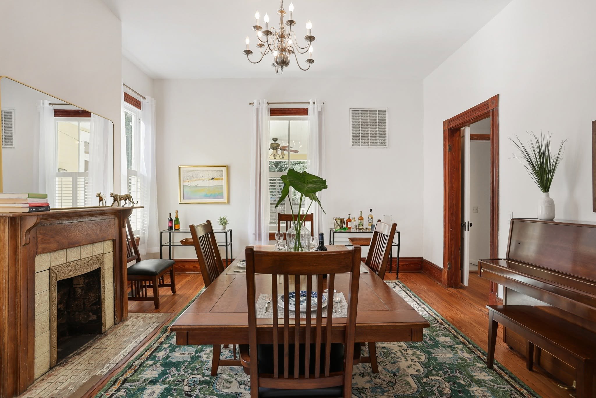 A dining room with wooden furniture, a fireplace, large windows with white curtains, a chandelier, and various decorative items including potted plants and framed artwork.