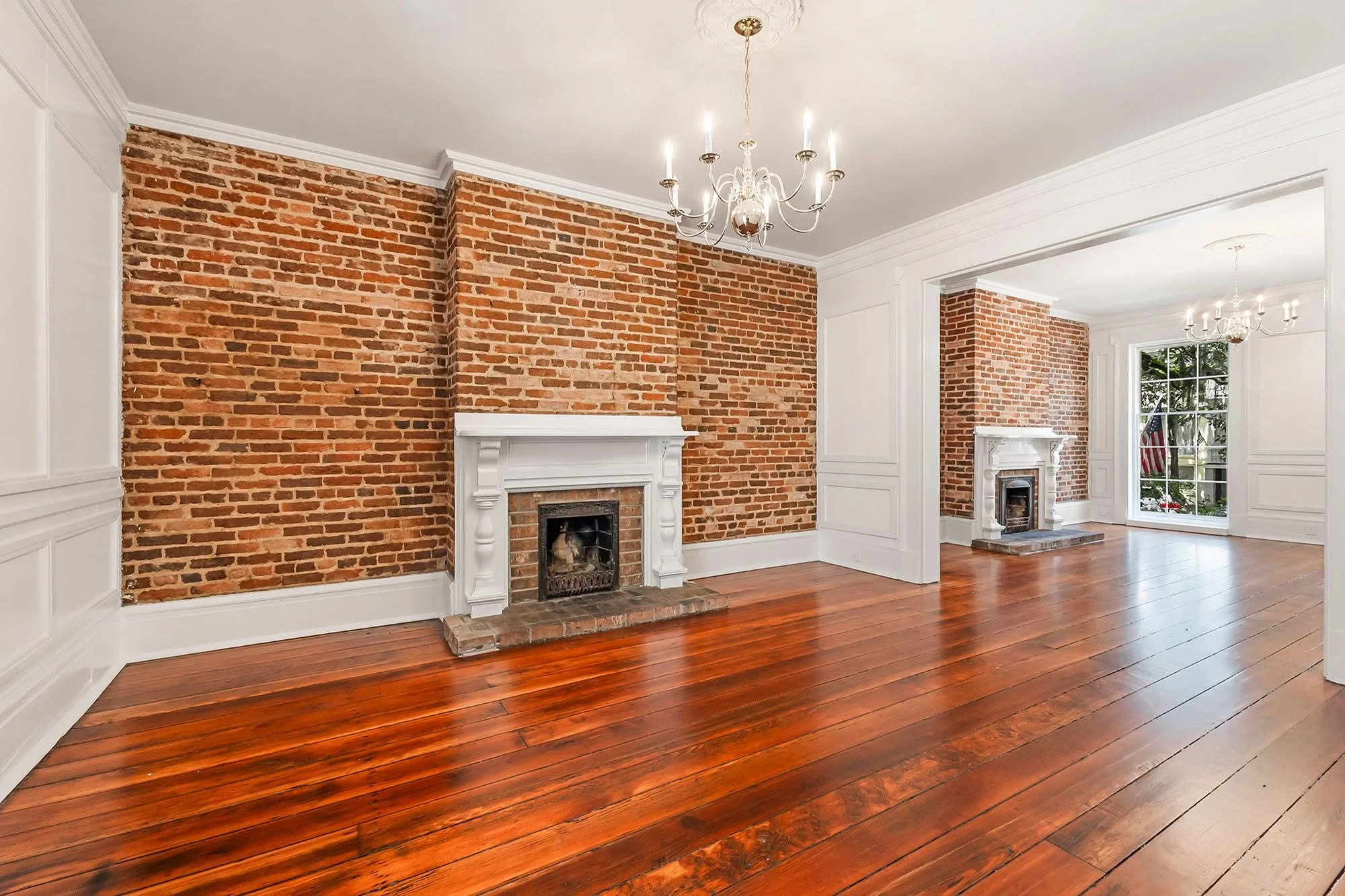 Living room with exposed brick walls, wooden floors, white decorative trim, and two white fireplaces with intricate mantels. Chandeliers hang from the ceiling, and sunlight streams through large windows, revealing an American flag outside.