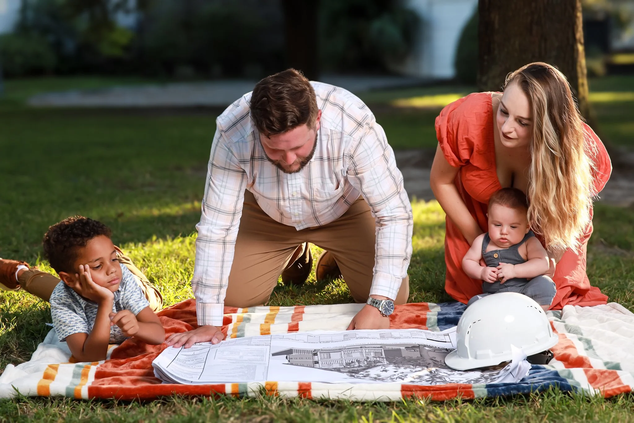 Group of adults and children on a grassy outdoor area, looking at blueprints or architectural plans on a blanket, with safety helmets nearby, in a park with trees and a waterfall in the background.