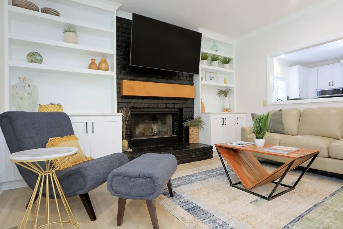Living room with a black brick fireplace, flat-screen TV above, white built-in shelves with decorative items, beige sectional sofa, gray armchair with ottoman, wooden coffee table with magazines, and a colorful area rug.