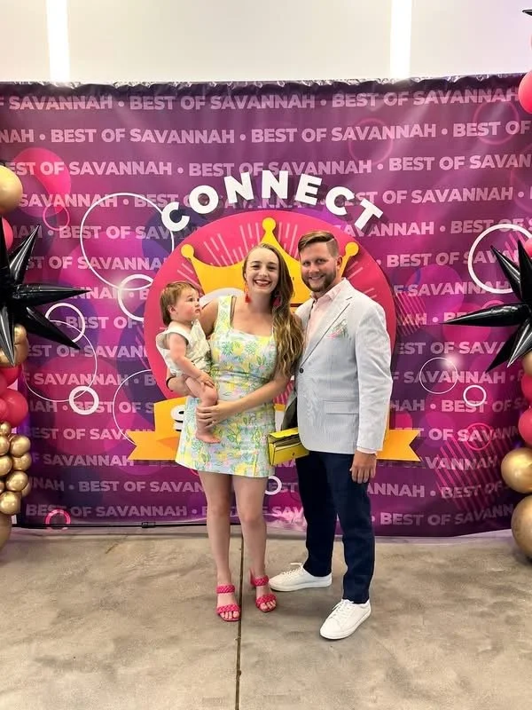 A woman holding a young girl, standing next to a man at an event with a pink and purple backdrop that reads 'Best of Savannah' and features a crown graphic and the word 'Connect'. Decorations include black and gold star-shaped balloons and gold balls.