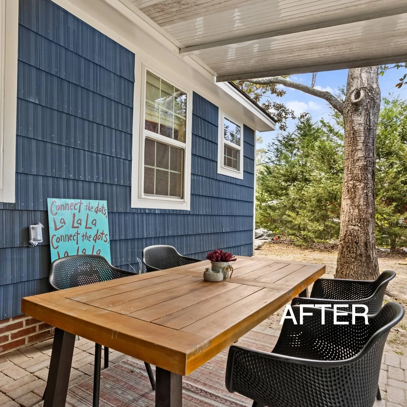 A backyard patio with a wooden table and black chairs. There is a blue house wall with two windows, a sign that says 'Connect the dots, La La La, Connect the dots, La La La.' A large tree and green bushes are in the background. The image is labeled '