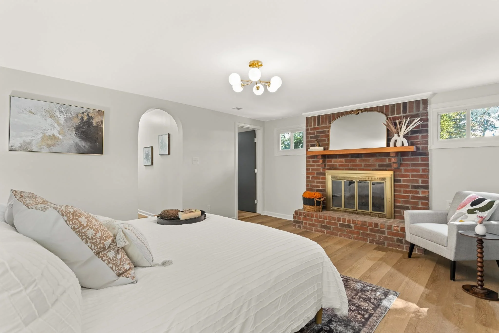 A cozy living room with a white couch, decorative pillows, a side chair, a brick fireplace with a mirror and vases on the mantle, and artwork on the wall. There are small windows near the ceiling, and a modern chandelier hangs from the ceiling.