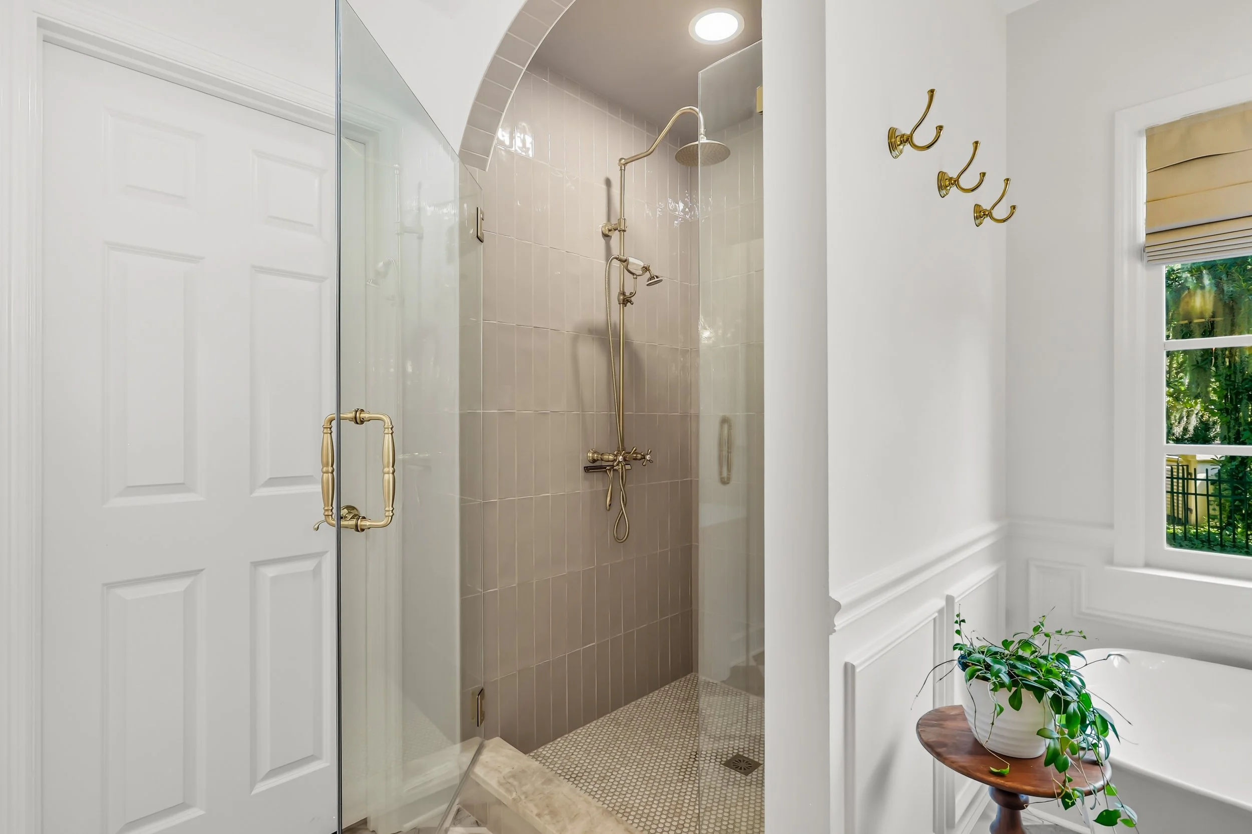 A bathroom with a walk-in shower that has gold fixtures, a glass door, and beige tiled walls. There is a window with a Roman shade, some hooks on the wall, and a small wooden table with a potted plant.