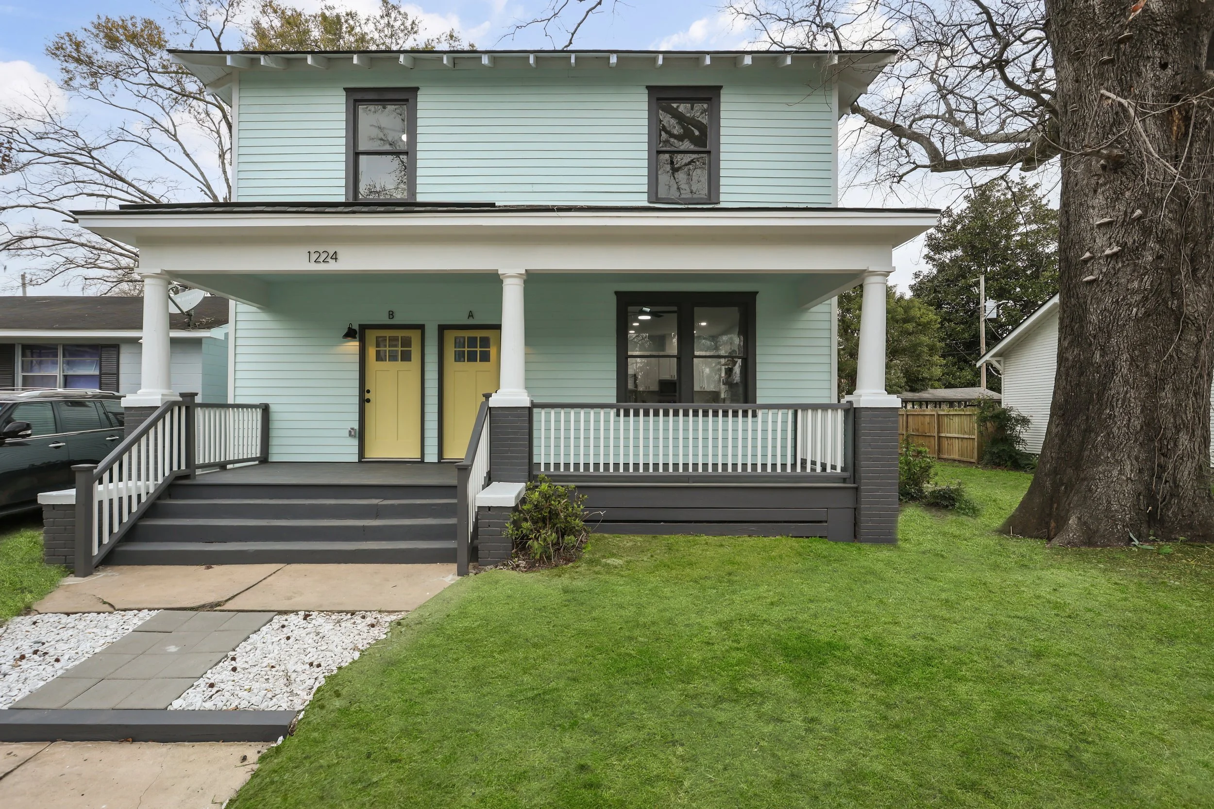 Front view of a two-story house painted in light blue with black window frames, yellow double front doors, and a porch with white and dark gray railings. A large tree is on the right side of the house, and the driveway has gray pavers with white deco