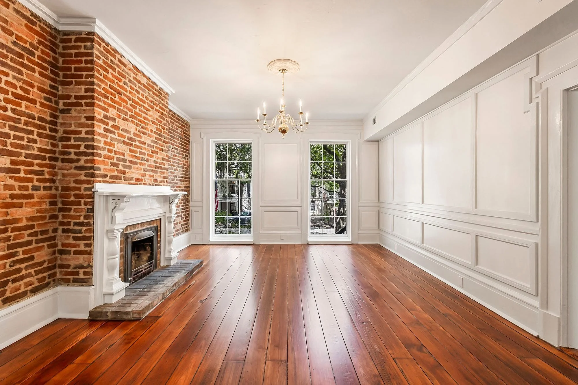 Living room with brick fireplace, hardwood flooring, white paneled walls, large windows, and a chandelier hanging from the ceiling.