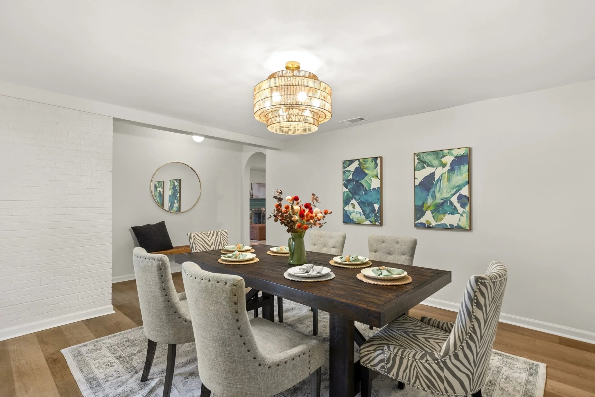 A dining room with a rectangular dark wood table set with six plates, napkins, and woven placemats, surrounded by upholstered chairs with different patterns, a vase of flowers, a wall mirror, colorful leaf artwork, a beige rug, a white brick wall, an