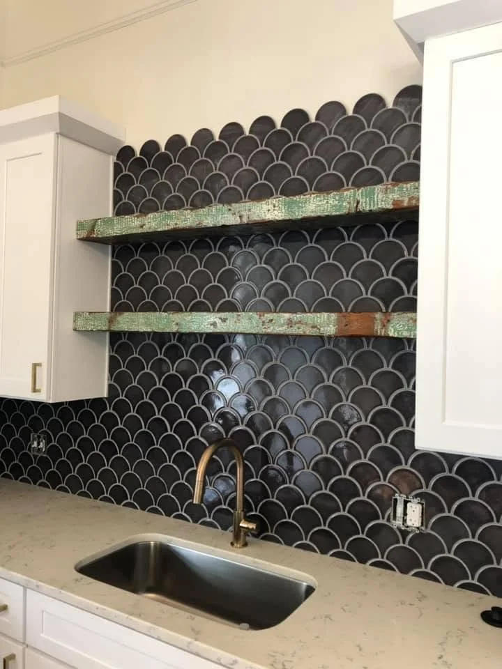 Black backsplash with a scallop tile pattern above a kitchen sink equipped with a brass faucet. Two wooden shelves, appearing weathered, are installed on the backsplash. White kitchen cabinets are on either side.