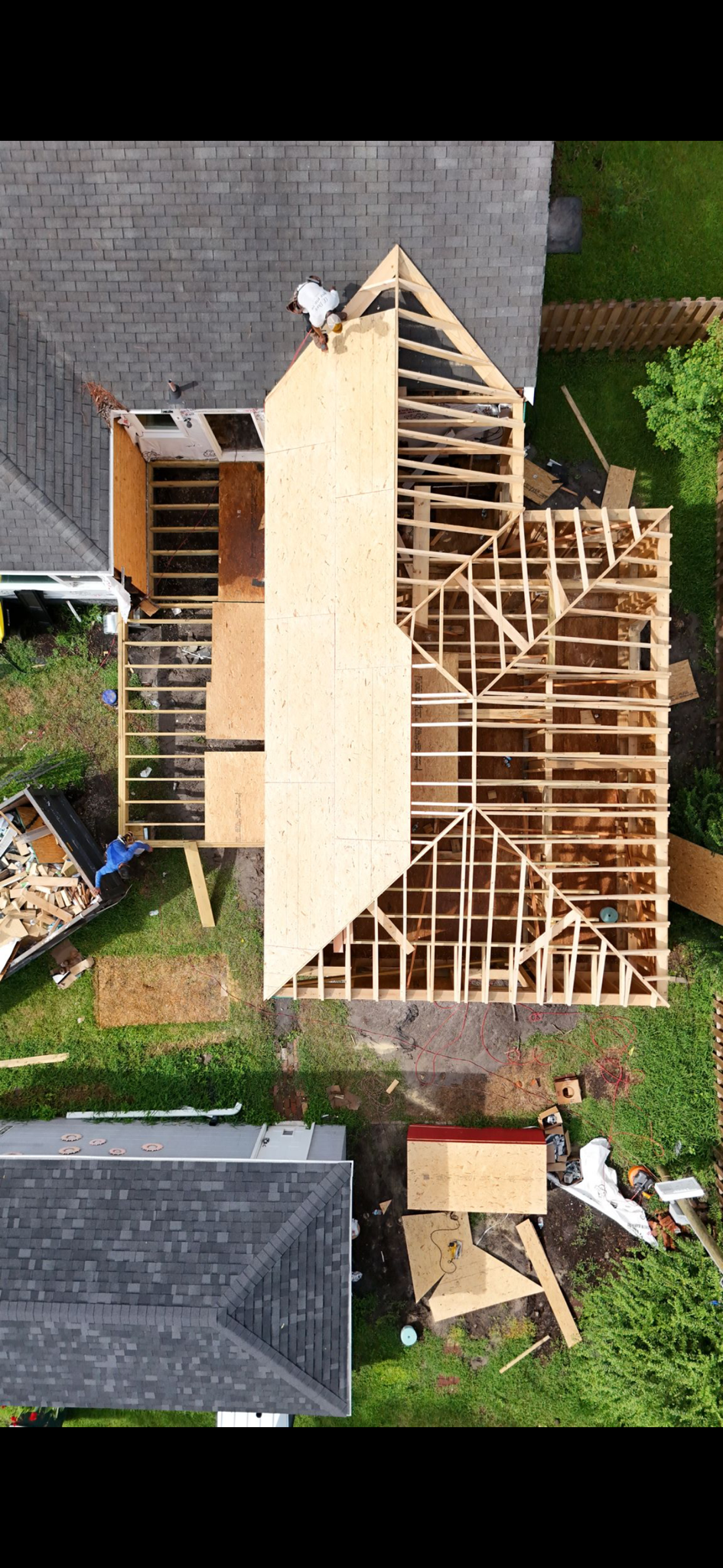 An aerial view of a house under construction with a roof framework and workers installing roof panels, surrounded by a yard with grass, a fence, and neighboring houses.
