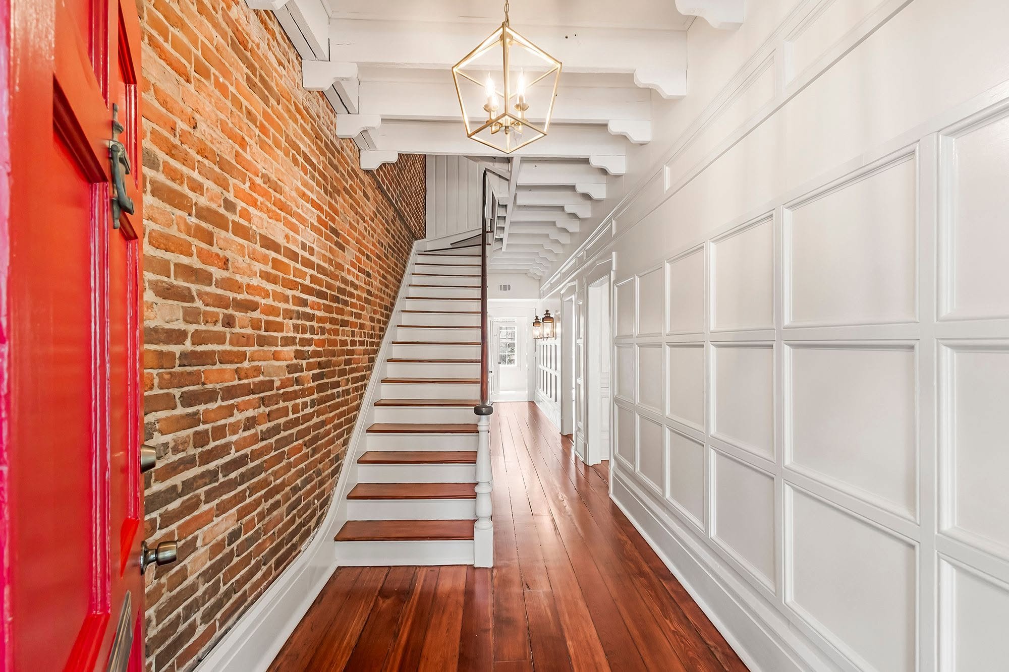 Interior view of a hallway with a brick wall on the left, white paneled wainscoting on the right, hardwood flooring, and a staircase with white risers and dark handrail leading upward. A modern geometric chandelier hangs from the ceiling.