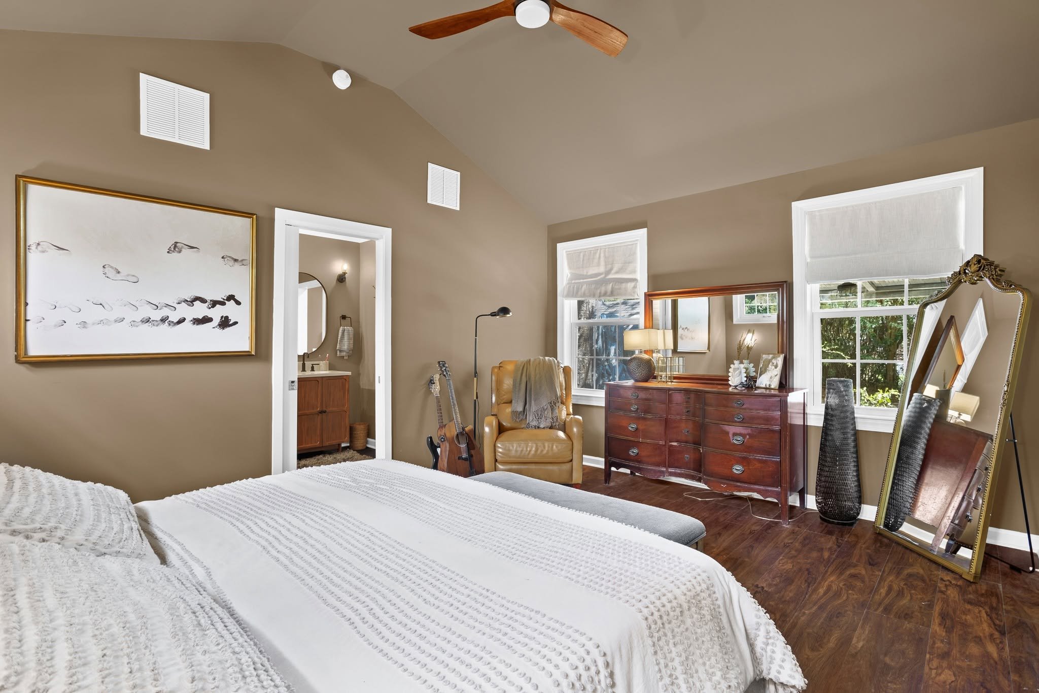 A cozy bedroom with a white textured bedspread, a wooden dresser topped with decorative items, a standing mirror, a yellow armchair, and musical instruments near the wall. The room has large windows with white blinds, a ceiling fan, and natural light