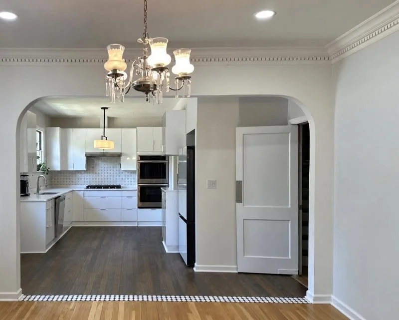 Modern kitchen with white cabinets, black appliances, and a wooden floor. There is a chandelier hanging in the dining area and a doorway with a sliding door on the right.