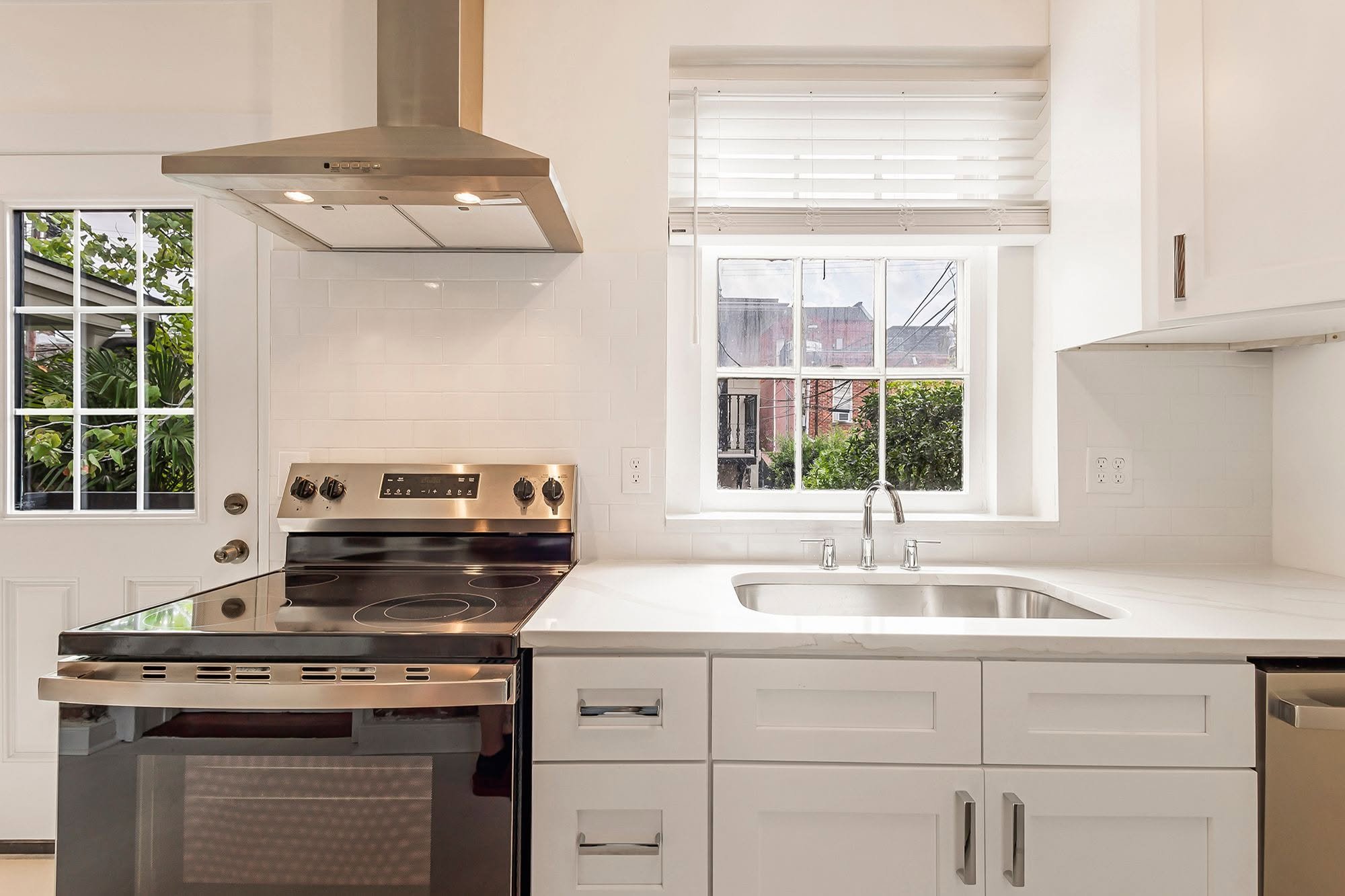 A modern kitchen with white cabinets, a white countertop, a stainless steel stove, a range hood, a double-basin stainless steel sink, and a window with horizontal blinds showing an outdoor view.