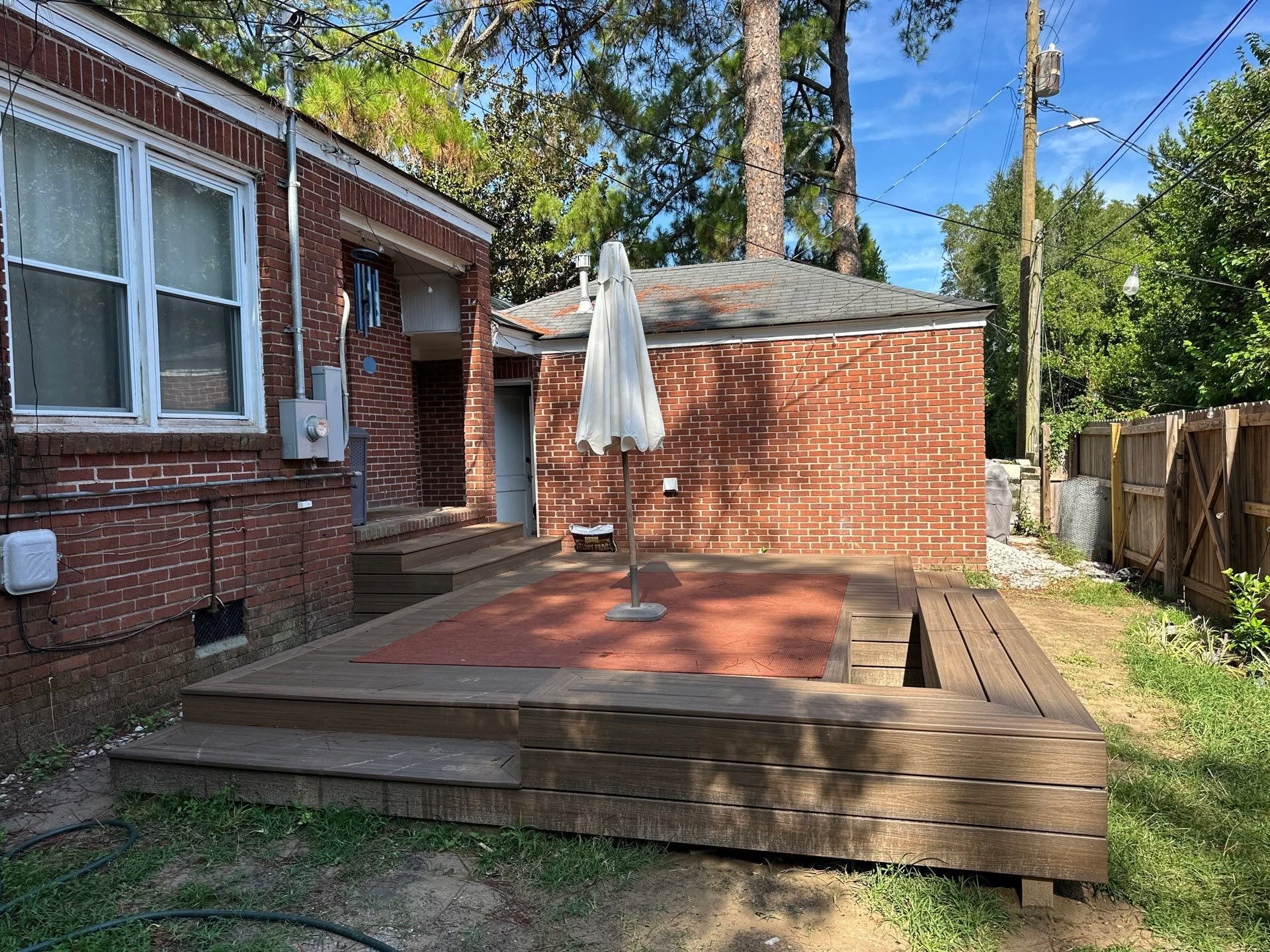 Backyard with newly built wooden deck, umbrella, brick house with patio, window, trees, power lines, and a clear blue sky.