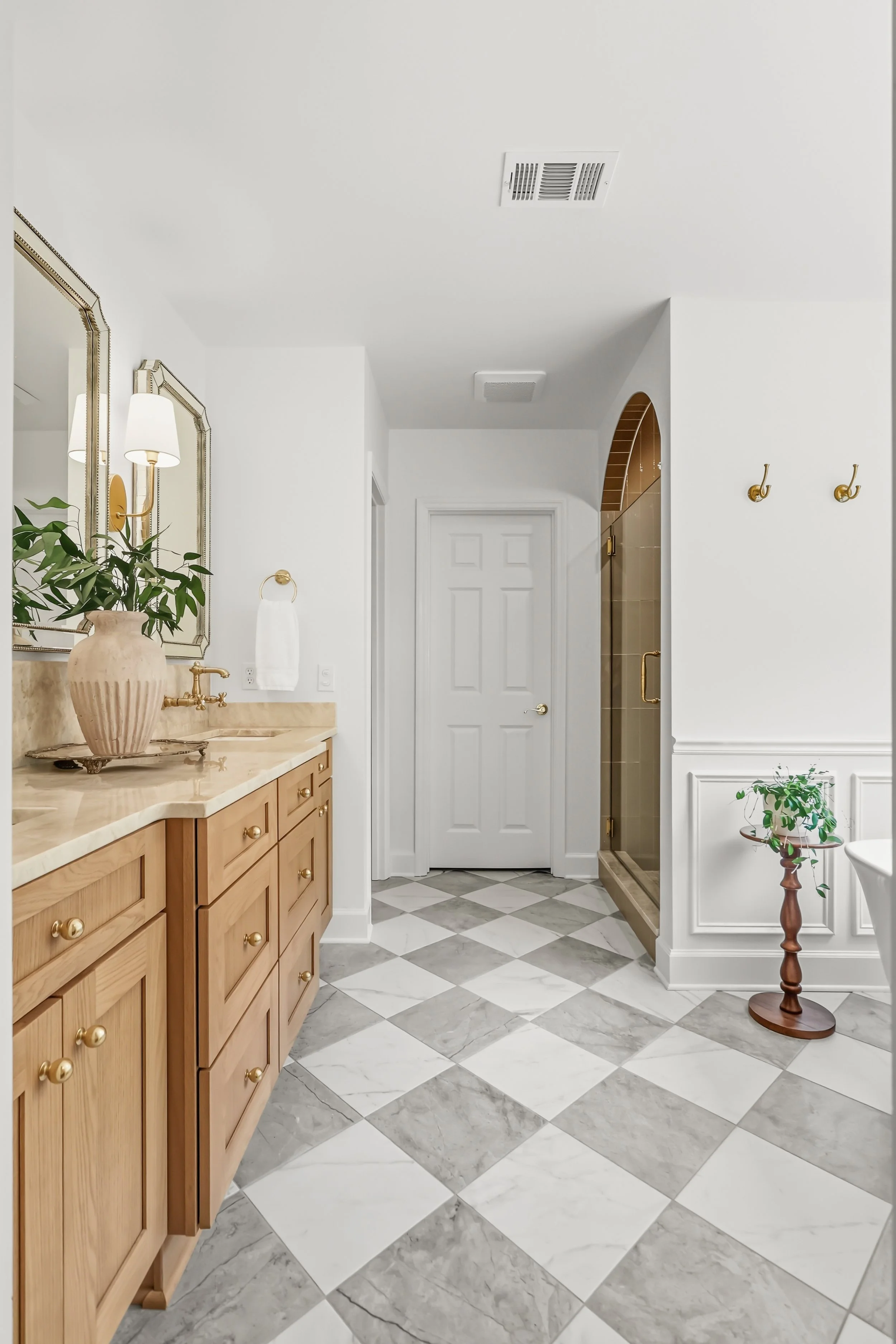 A bathroom with a wooden vanity with a marble countertop, a large mirror, and a glass-enclosed shower. There are gold fixtures and wall hooks, a potted plant, a white towel, and decorative elements like a beige vase and a small table.
