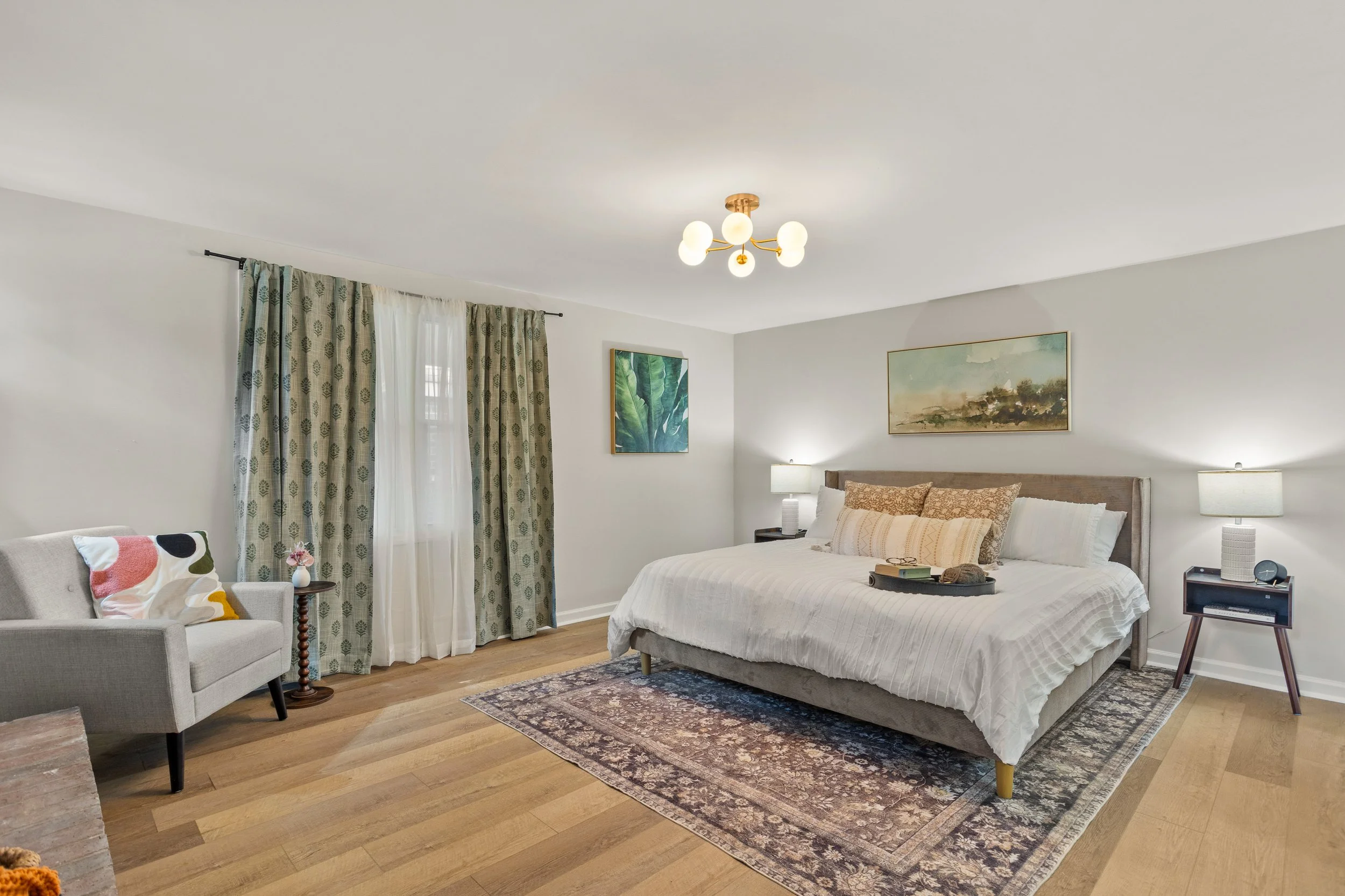 A cozy bedroom with a large bed, two nightstands with lamps, framed artwork on the wall, a window with curtains, a sitting area with a sofa and a side table, and a patterned rug on hardwood floor.