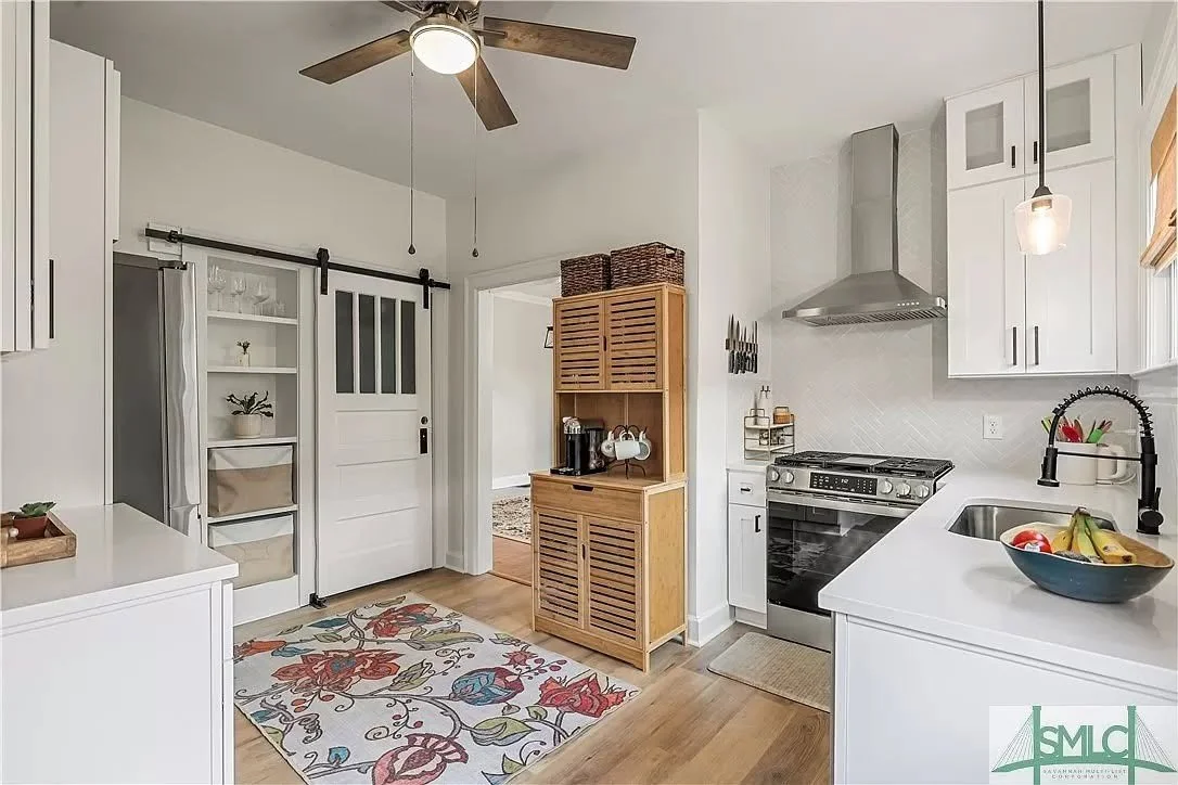 A kitchen with white cabinets, a stainless steel stove and range hood, a white farmhouse sink, a wooden rolling butcher block, colorful floral rug, and a ceiling fan with wooden blades.