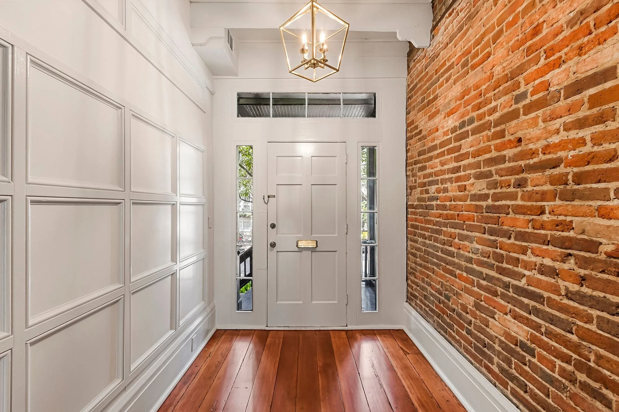 Entryway with white paneled walls, a brick accent wall, wooden floor, white front door with glass panels, and a geometric gold chandelier.