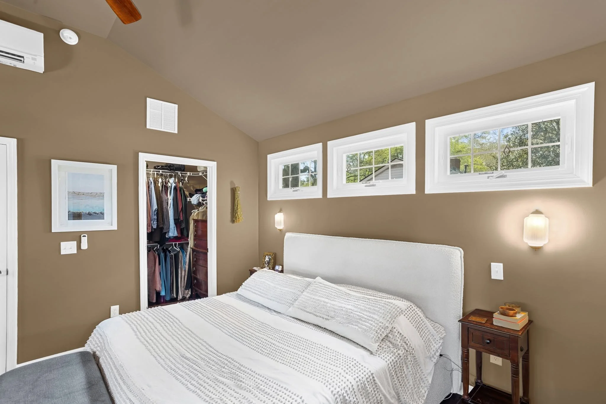 A bedroom with a white bed, wooden nightstand, beige walls, and three rectangular windows letting in natural light. An open closet with clothes is visible, and a framed picture hangs on the wall.