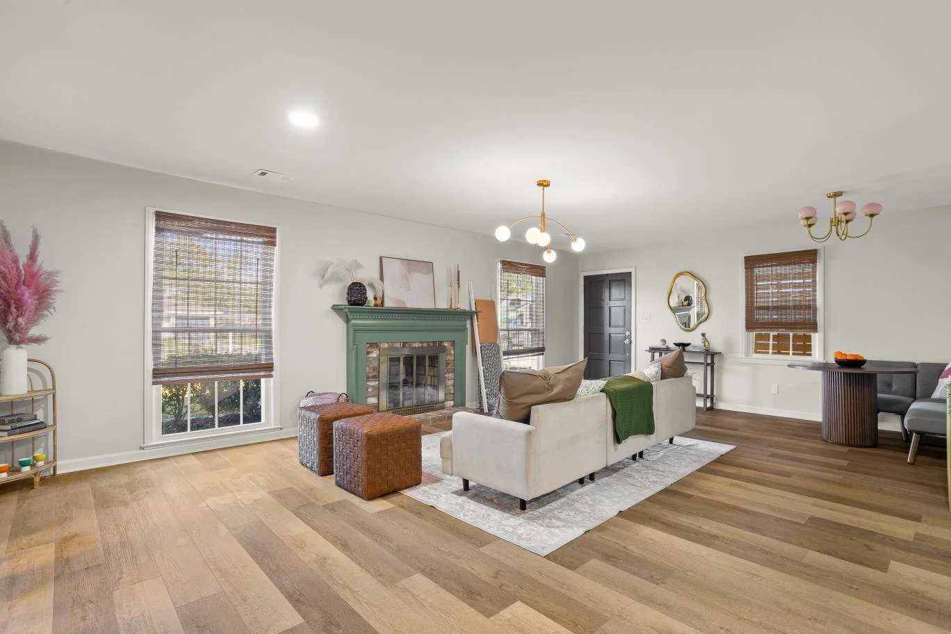 Spacious living room with hardwood floors, white walls, a fireplace with a teal mantel, a cream sofa with brown and white pillows, two brown ottomans, a modern chandelier, and multiple windows with wooden blinds.