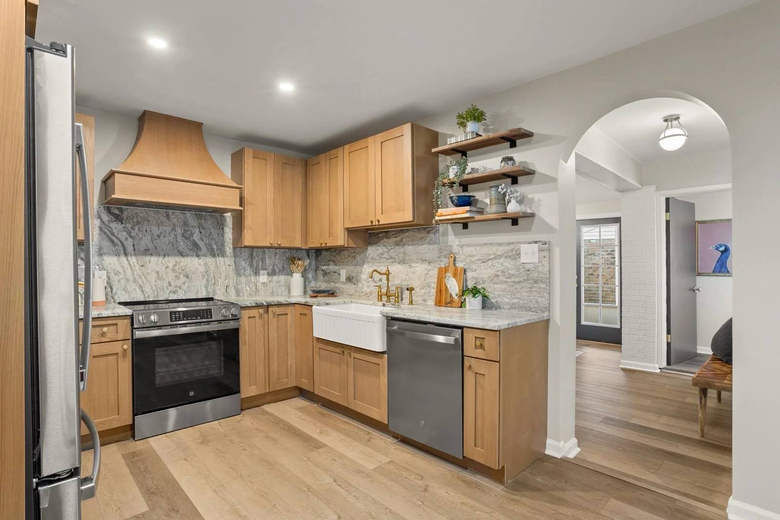Kitchen with wooden cabinets, gray marble backsplash, stainless steel appliances, a farmhouse sink, open shelving with plants and dishes, and a doorway leading to a hallway with a bench and artwork.