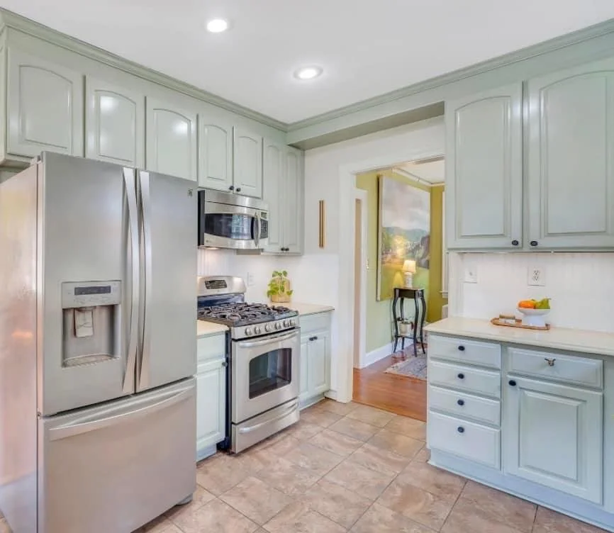 A kitchen with light blue cabinets, stainless steel refrigerator, microwave, and stove, beige tile floor, and doorway leading to a room with wooden floor and a painting.
