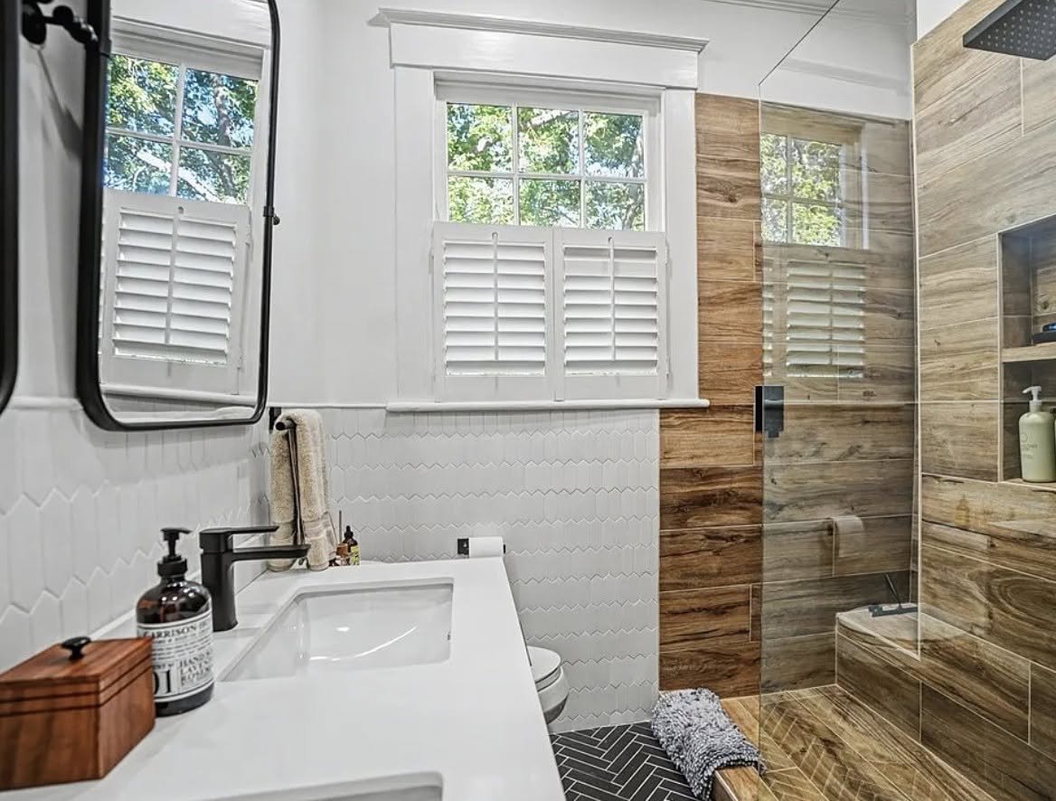 Modern bathroom with a shower area featuring wood-like tiles, white wall tiles, a white vanity with a sink, a mirror, and windows with white shutters.
