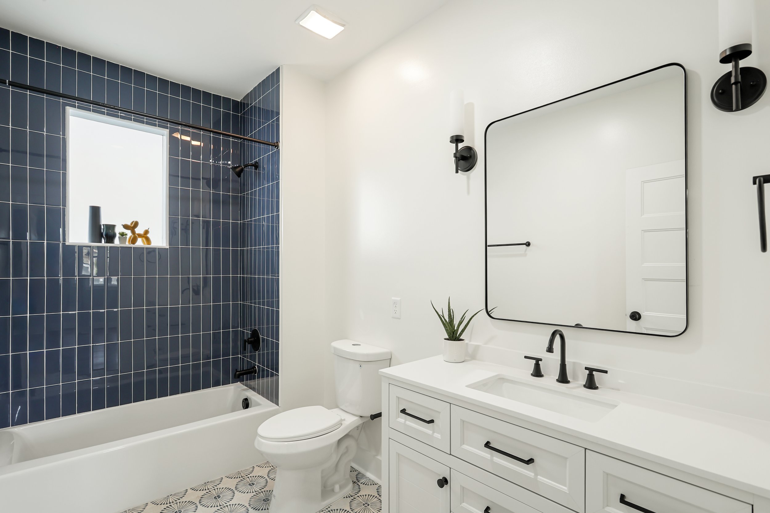 A modern bathroom with a white vanity, black fixtures, a large mirror, and a blue-tiled shower area with a window and black shower fixtures.