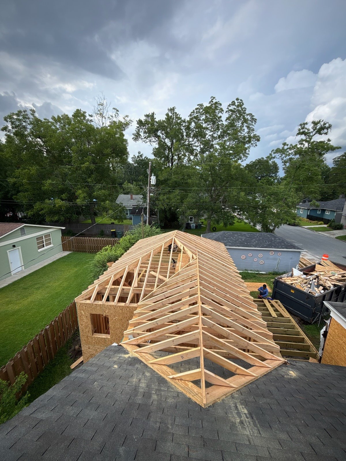 A house under construction with a wooden roof frame, surrounded by a fenced backyard and neighboring houses, with a cloudy sky overhead.