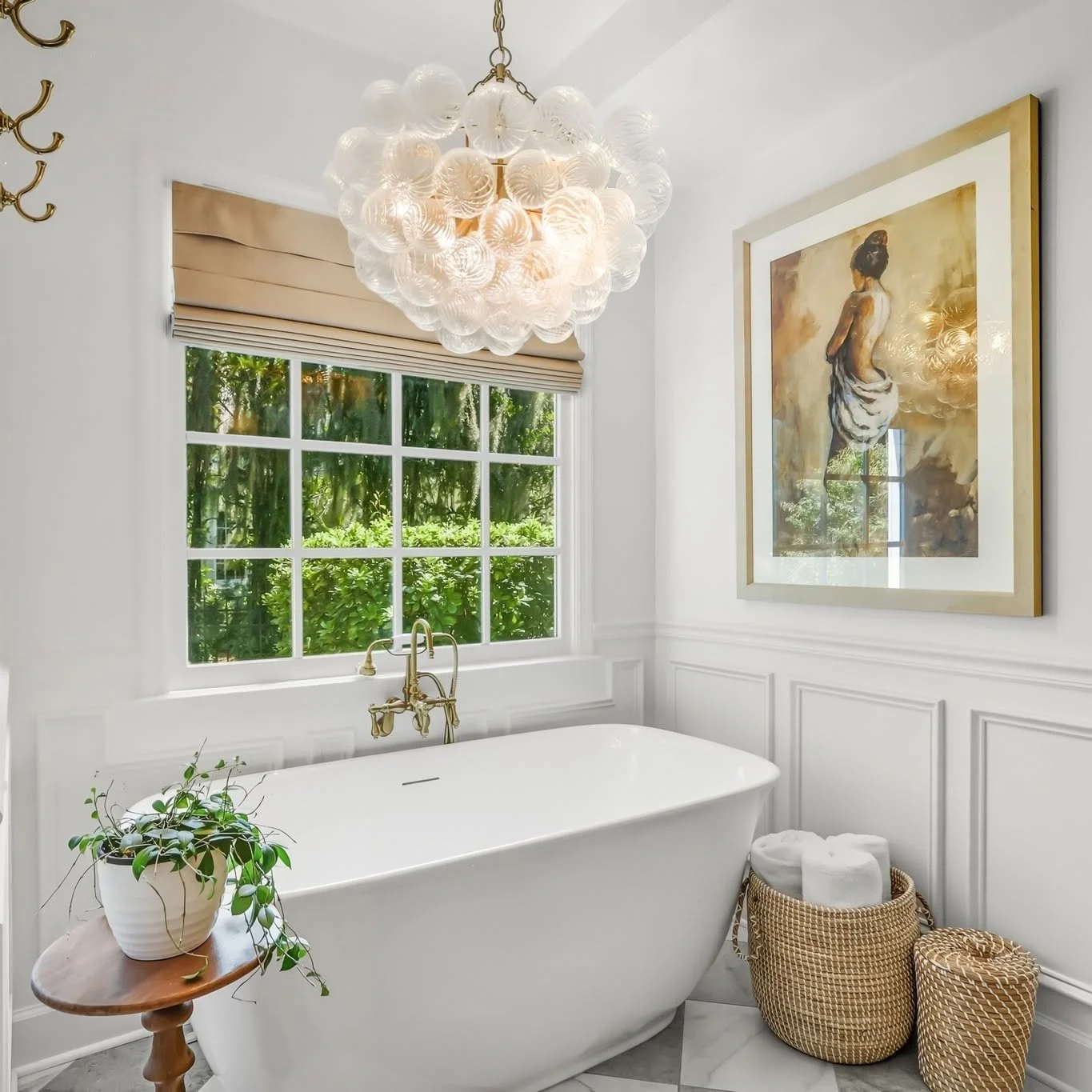 Bright bathroom with a white freestanding bathtub, a brass faucet, a window with a beige Roman shade, a chandelier made of glass globes hanging from the ceiling, a framed artwork of a woman, a potted plant on a wooden table, and woven baskets with rolled white towels.
