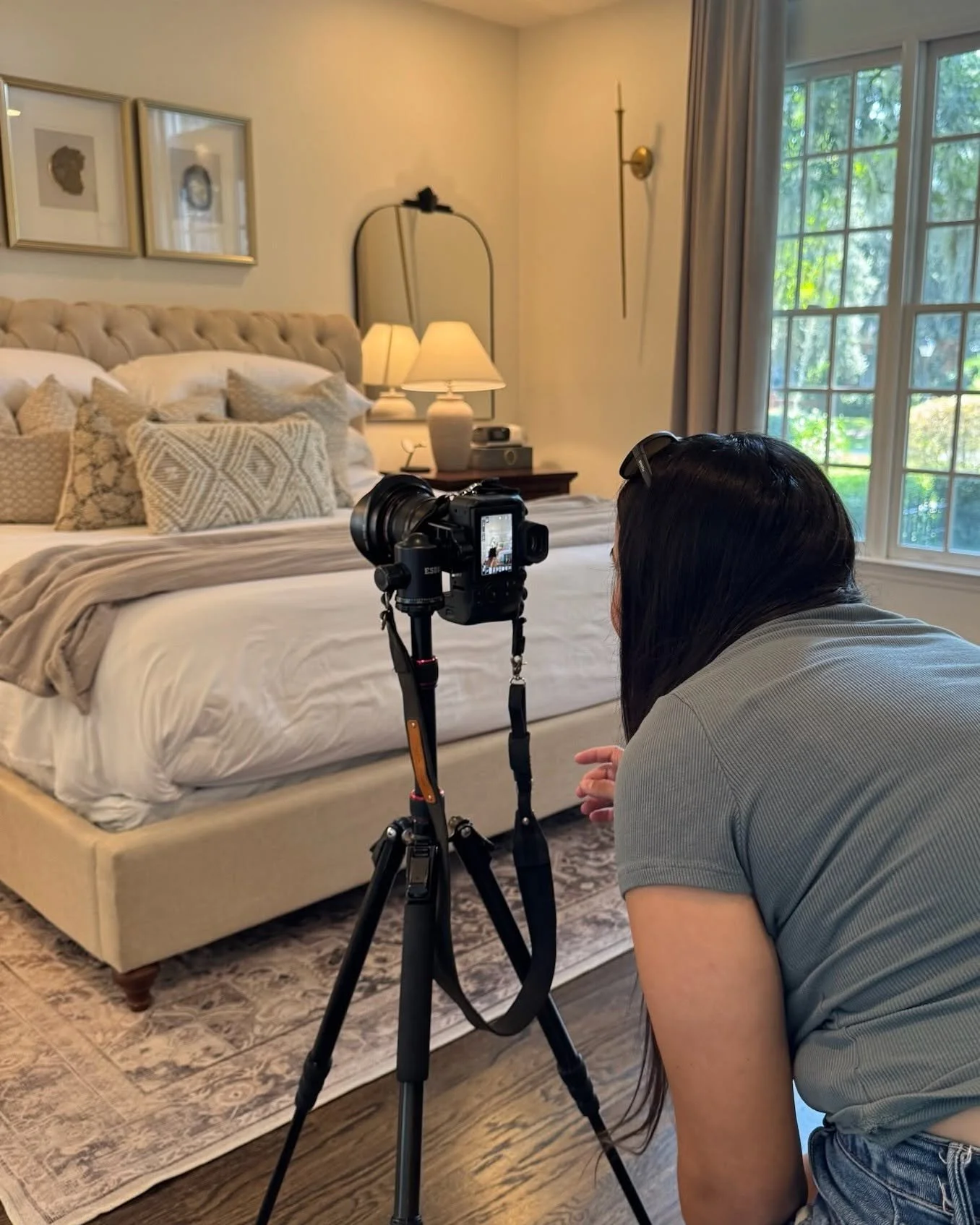A woman with dark hair, wearing a gray shirt and glasses on her head, is kneeling behind a camera on a tripod, capturing a photo or video of a neatly made bed in a bedroom with a large window, curtains, bedside lamps, and wall art.