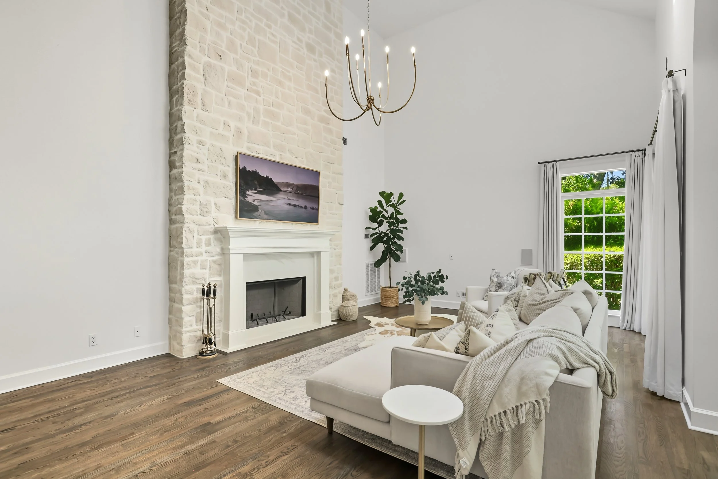 Interior view of a living room with white walls, a beige sofa with pillows, a white fireplace with a landscape painting above, a brass chandelier, a potted plant, and a window with white curtains showing green trees outside.
