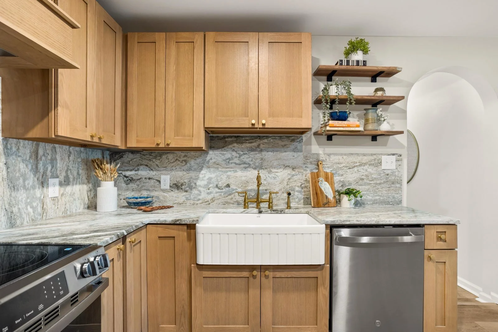 Kitchen with wooden cabinets, granite countertops, a white farmhouse sink, a dishwasher, and open wooden shelves with decorative items.