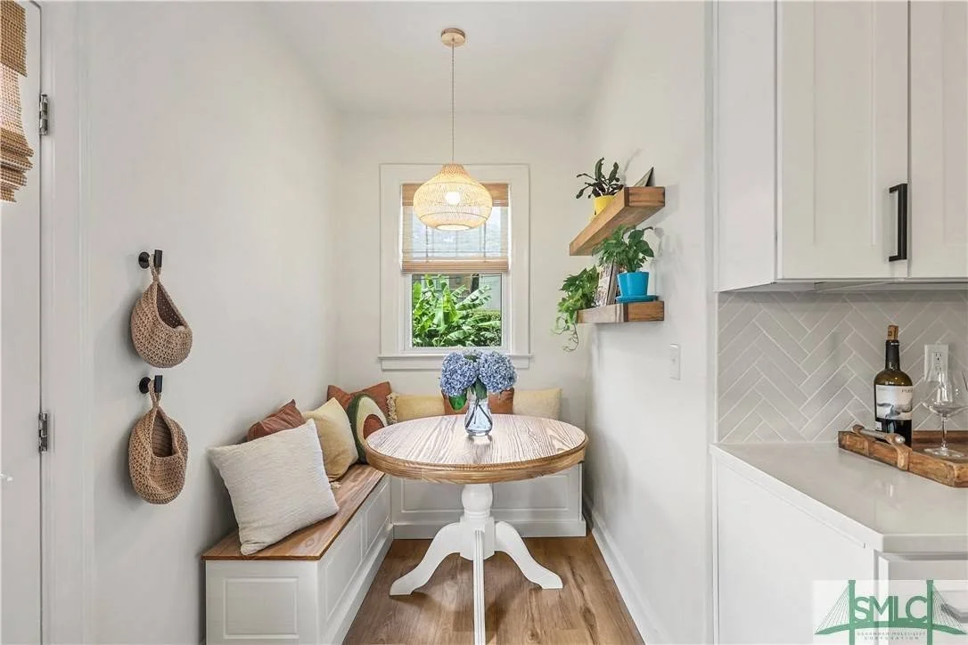 A small dining nook with a round wooden table, a bench with throw pillows, a window with blinds, and wall shelves with plants. There are two woven baskets hanging on hooks on the wall and a light fixture hanging from the ceiling.