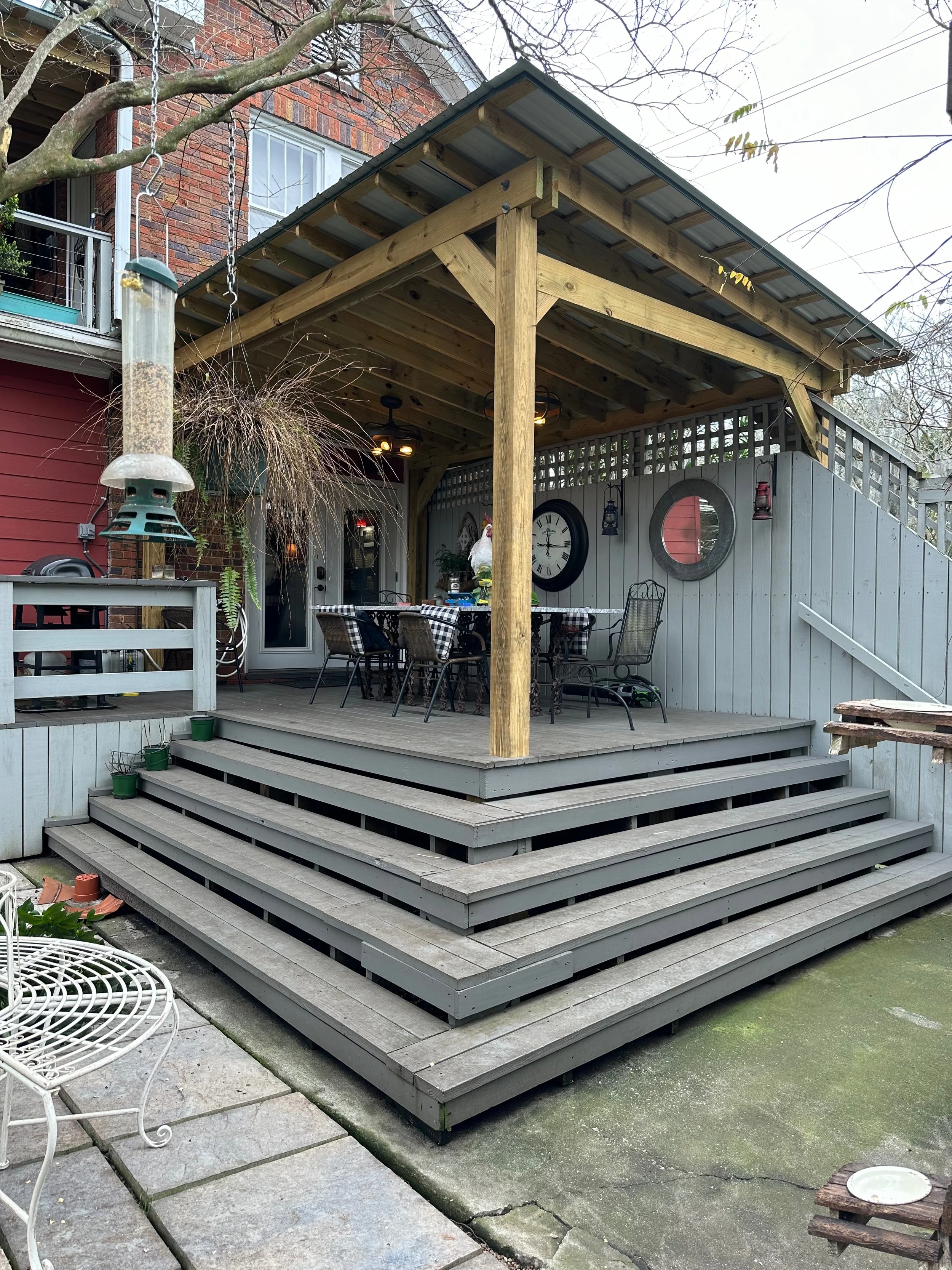 Backyard patio with a wooden deck and stairs, outdoor furniture, a large wall clock, and various decorative items including a chicken figure and hanging lanterns. There is a tree with a bird feeder and a small white wire chair on the patio.