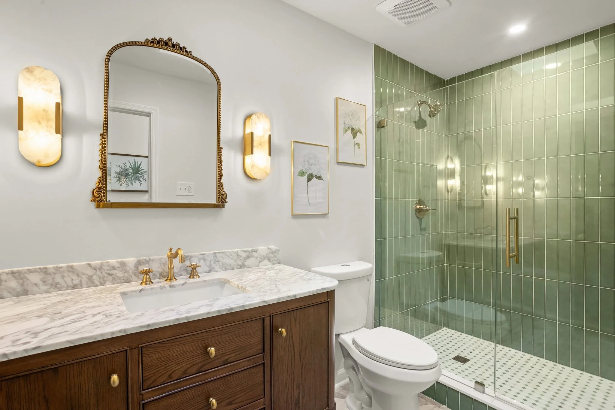 A bathroom featuring a green-tiled shower area with a glass door, a wooden vanity with a marble countertop, a white toilet, and decorative elements including framed botanical prints and wall-mounted light fixtures.