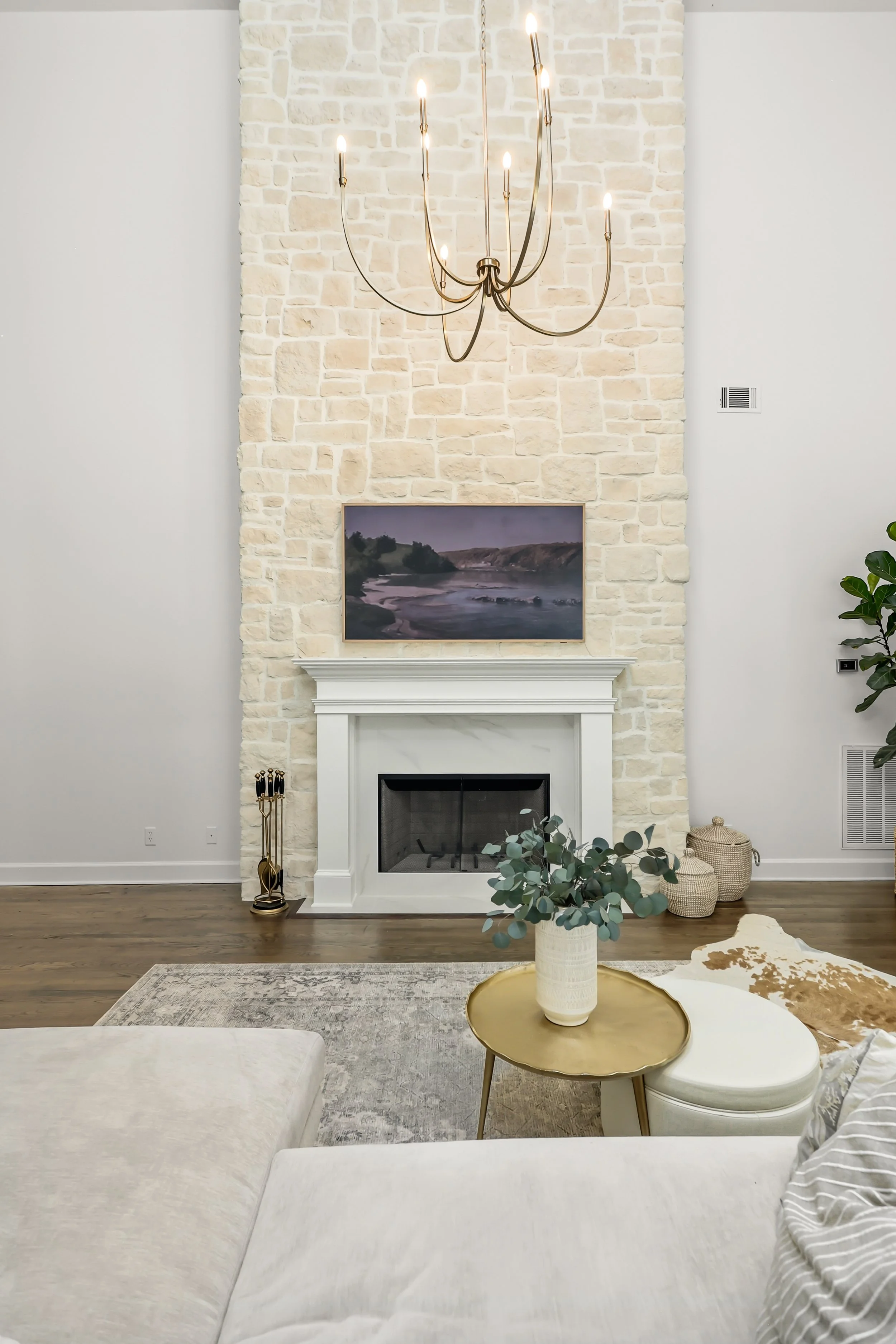 Living room with a white fireplace, a stone accent wall, a modern chandelier, a landscape painting above the fireplace, a cream-colored sofa, a round gold tray table with a white vase and eucalyptus, and woven baskets on the floor.