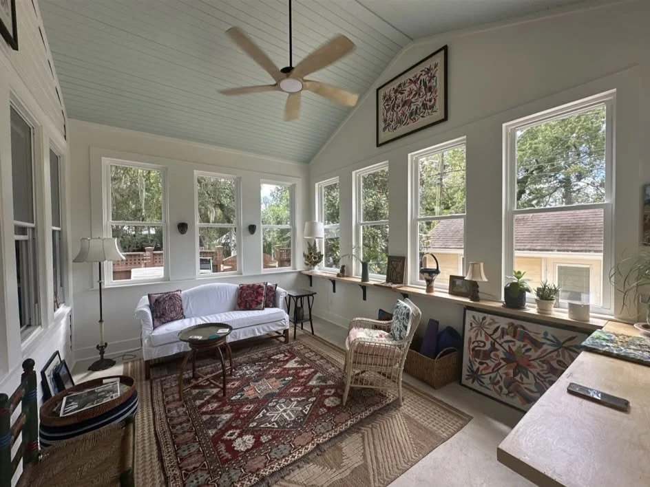 Bright sunroom with white walls and multiple windows, featuring a ceiling fan, white sofa with pillows, patterned area rugs, wicker chairs, and decorative objects like lamps, plants, and framed art.
