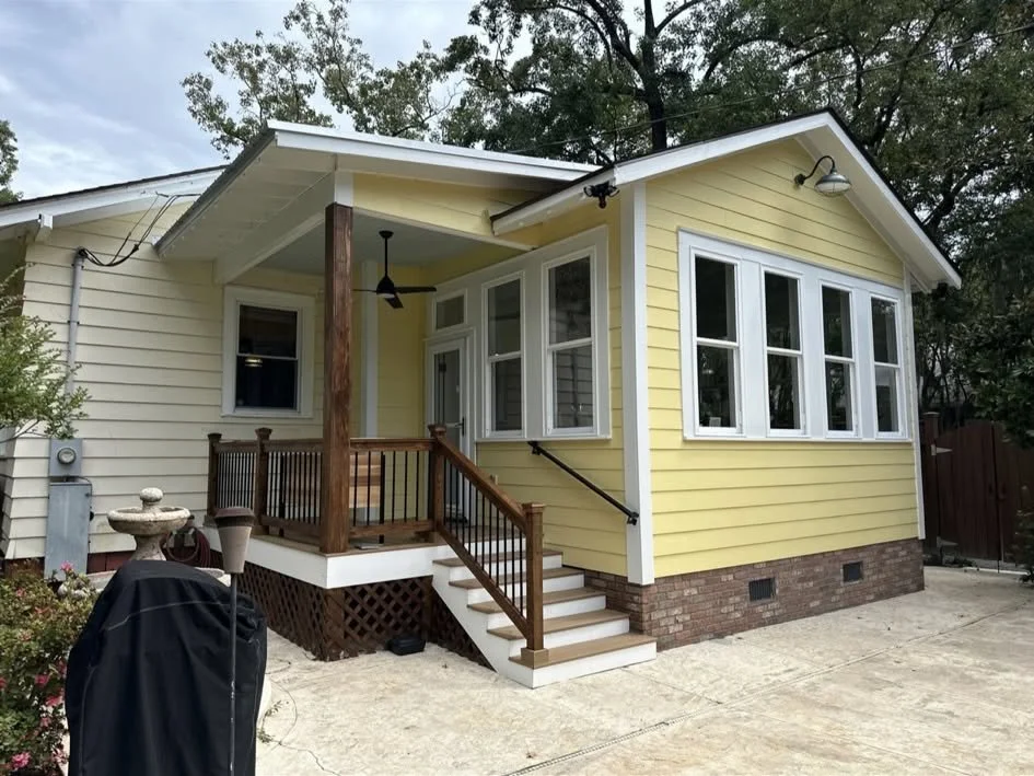 A small yellow house with white trim and a brick foundation, featuring a porch with wooden railings and stairs, surrounded by an outdoor concrete area and trees in the background.
