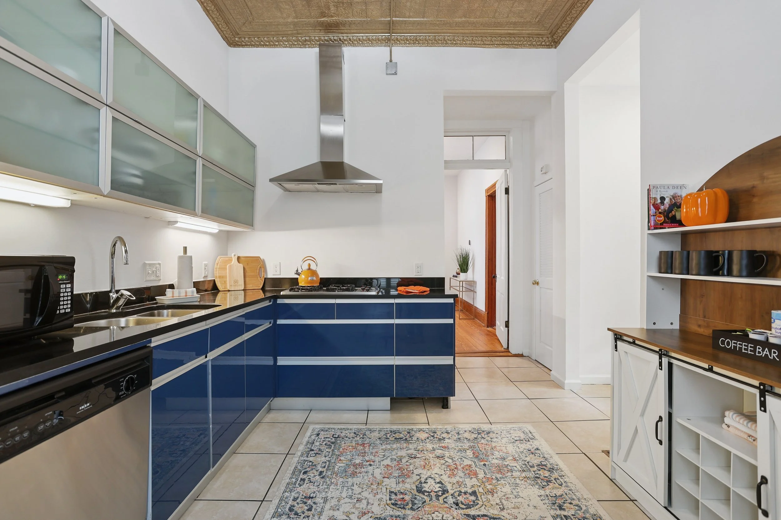 Modern kitchen with blue cabinets, stainless steel range hood, white walls, wooden accents, area rug, and open shelving with various items.