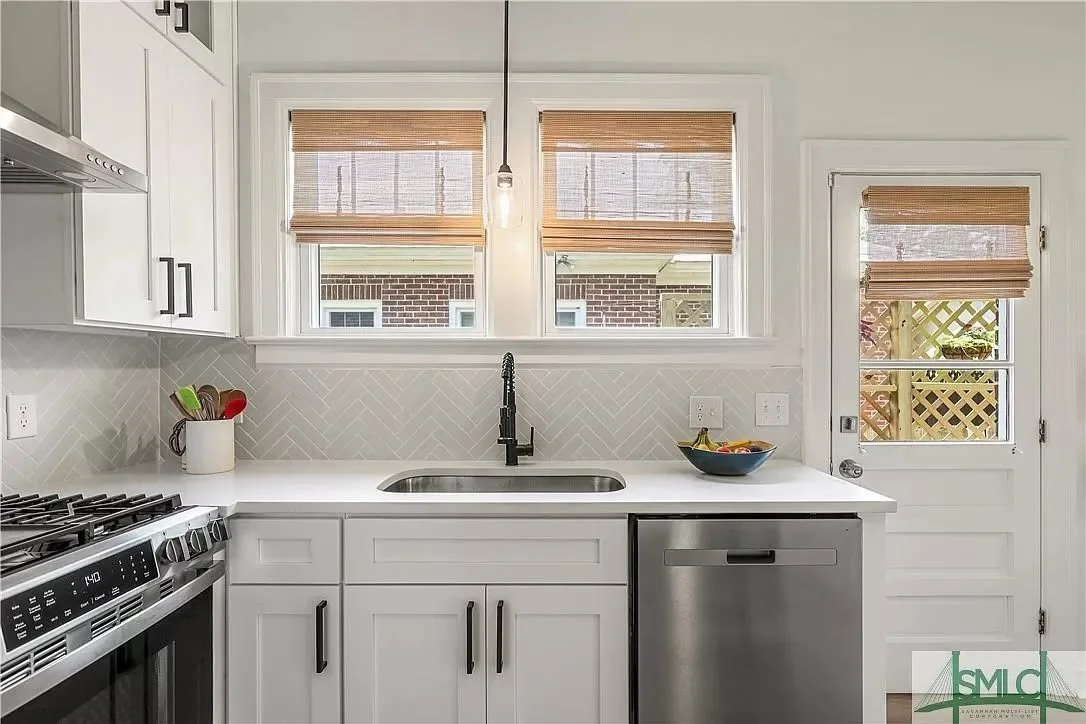 White kitchen with three windows above the sink, beige blinds, stainless steel dishwasher, black faucet, and white cabinets with black handles.