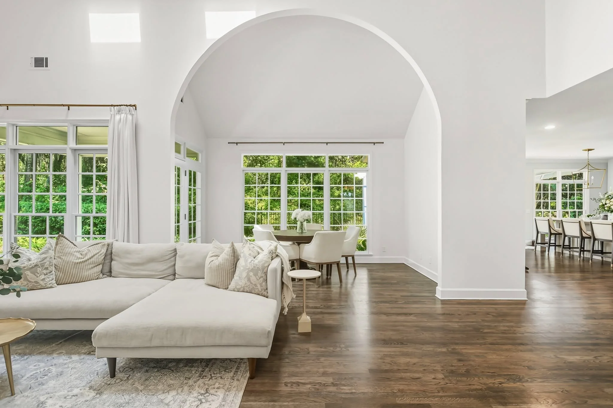 Bright and spacious living room with large windows, white walls, high vaulted ceiling, white sofa with striped pillows, round side table, and hardwood floors, overlooking a lush green outdoor garden.