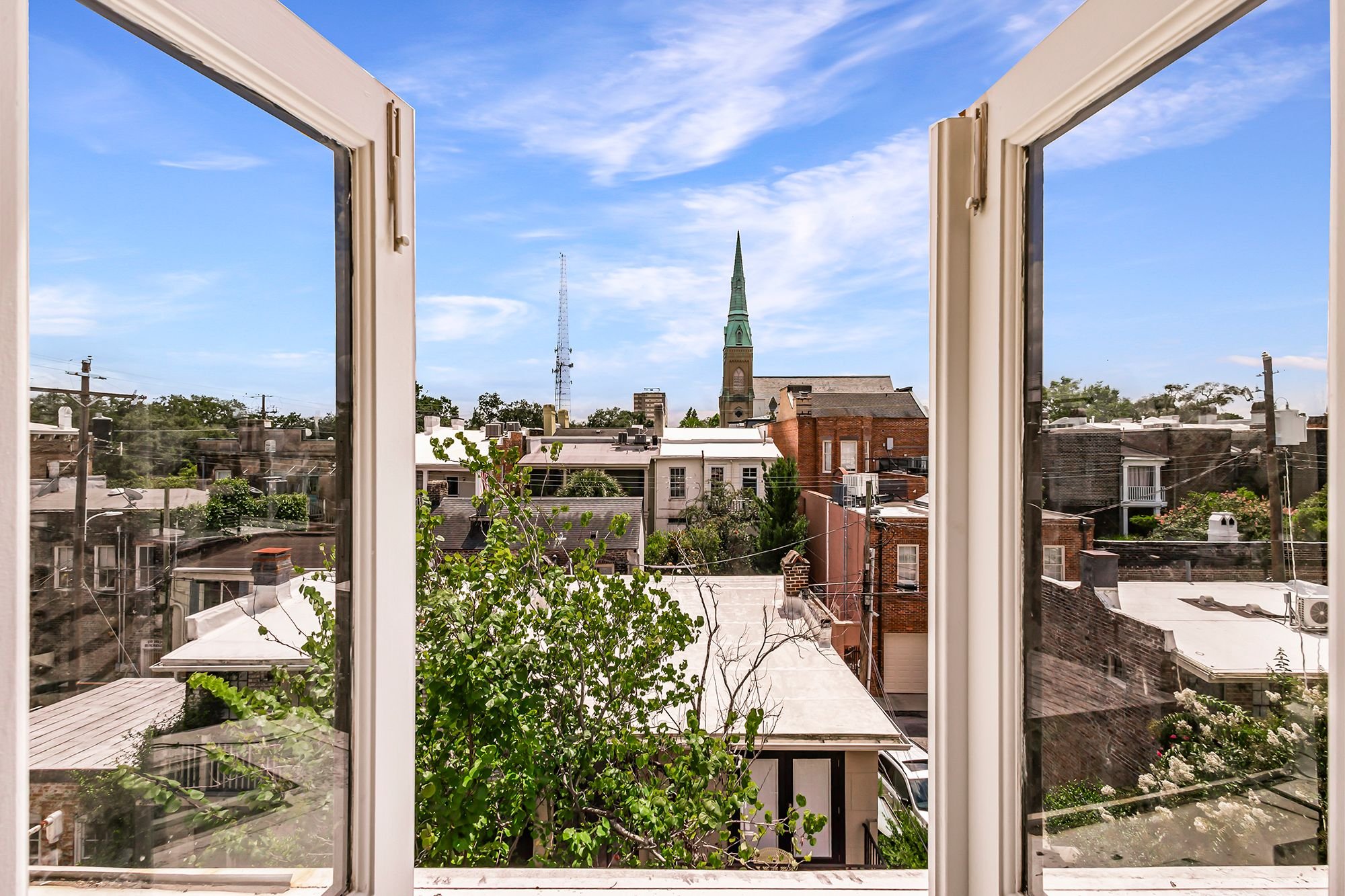 Looking through an open window at a cityscape with a church steeple, rooftops, trees, utility poles, and a blue sky with scattered clouds.