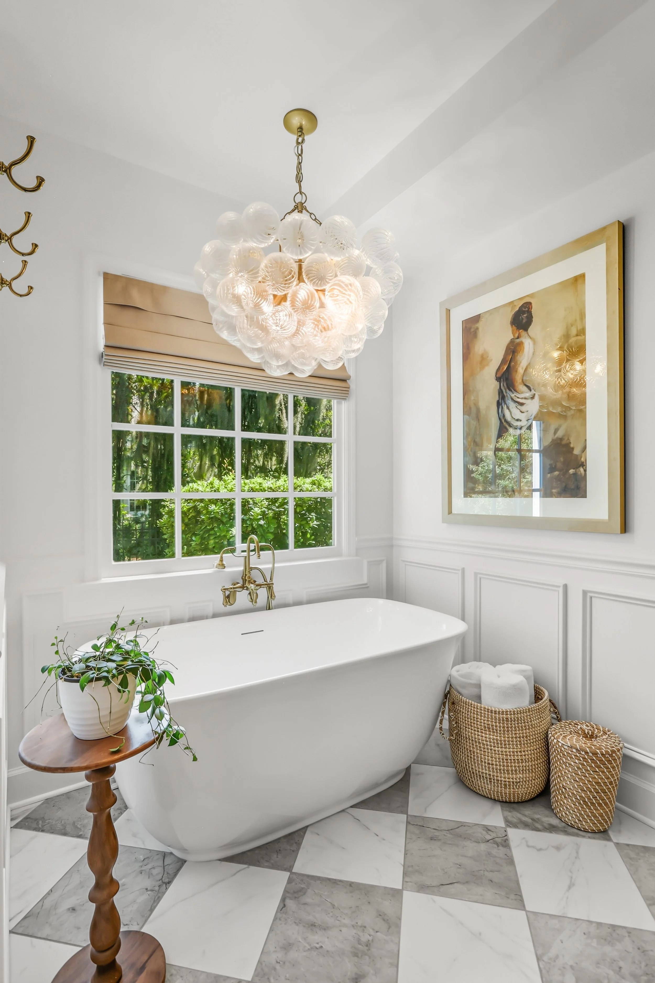 A bathroom with a white bathtub, a wooden side table with a potted plant, a gold faucet, a large window with beige blinds, a framed artwork of a woman, a modern chandelier with glass globes, and two woven baskets with towels, all with a marble floor 