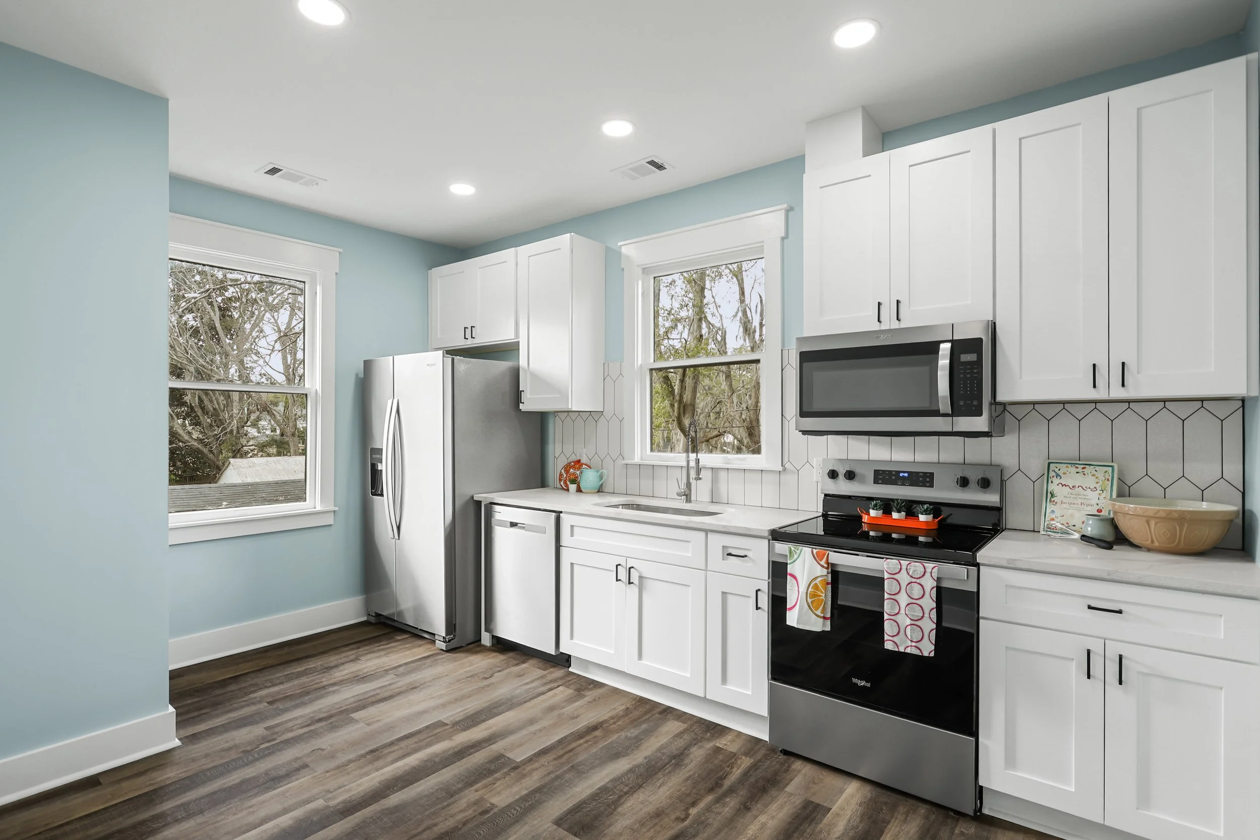 Kitchen with white cabinets, stainless steel appliances, light blue walls, two large windows, wood-look flooring, and ceiling lights.