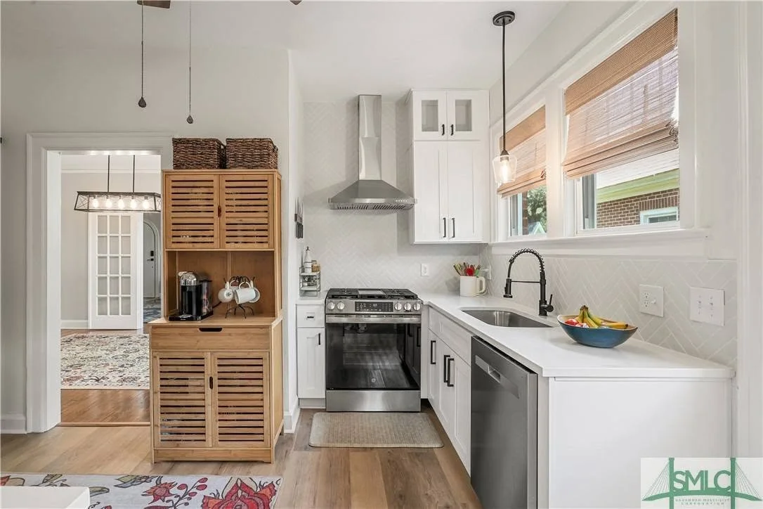 Kitchen with white cabinets, stainless steel appliances, black faucet, and light wood flooring. There are three windows with blinds, and a dark wood cabinet with baskets on top is visible on the left.